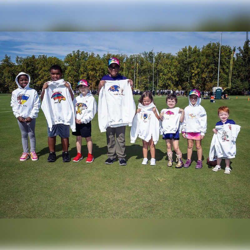 A group of children and an adult hold up white hoodies on a green grass field with a line of trees in the background.