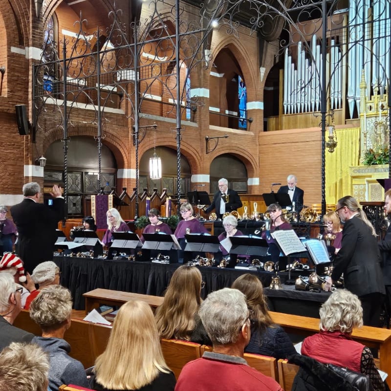 An orchestra performs on a stage in a grand brick cathedral, with an audience seated in pews in the foreground.