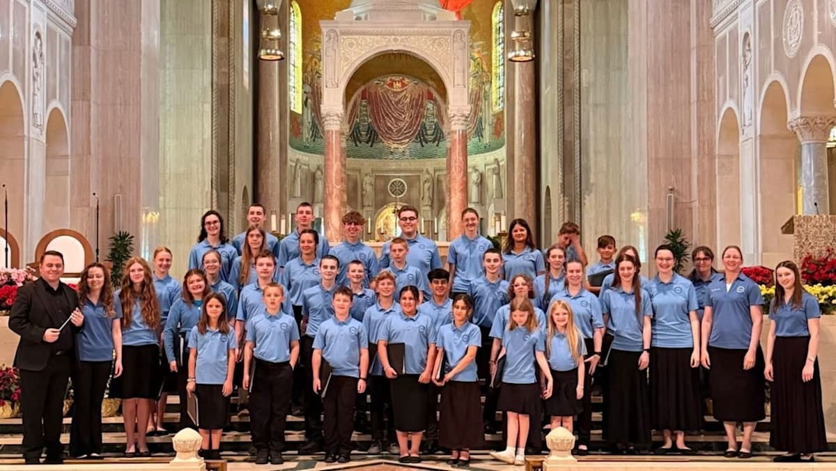 A large choir of students in blue shirts and black bottoms poses for a group photo inside a grand cathedral with ornate architectural details.
