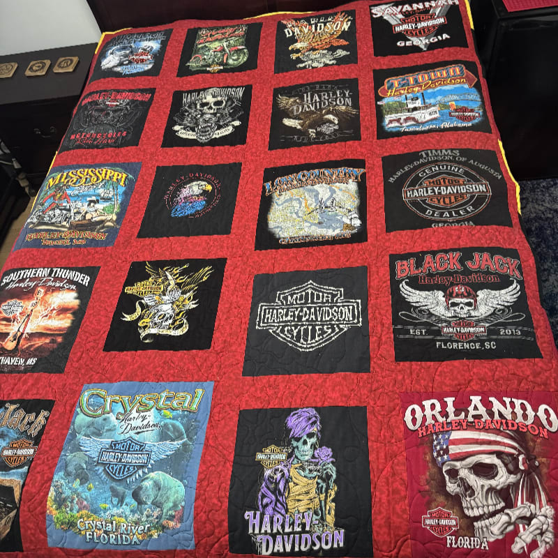 A red quilted blanket features a grid of various Harley-Davidson patches, resting atop a dark piece of furniture.