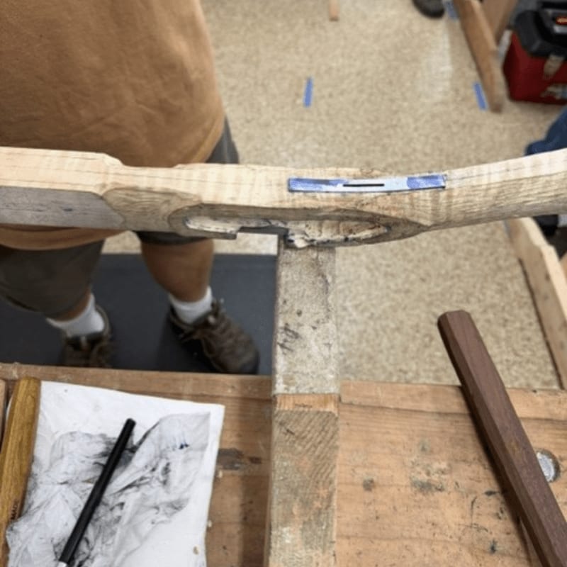A carved wooden stock rests on a workbench in the foreground, with a person standing in a workshop in the background.