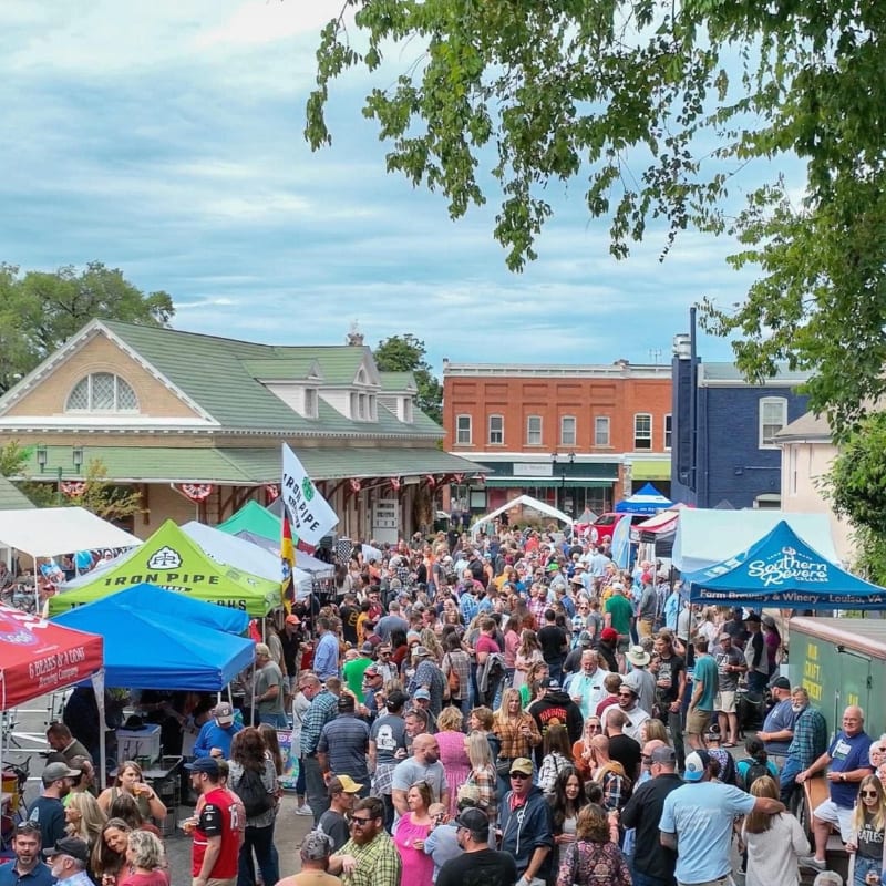 A crowded outdoor market with colorful tents in the foreground and buildings under a cloudy sky in the background.