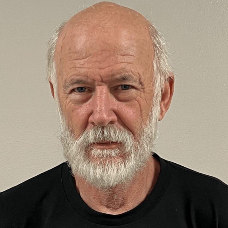 An older man with a white beard and a black shirt stands against a plain, light-colored wall.