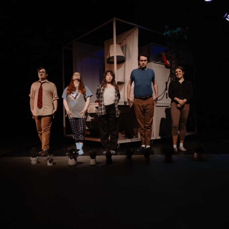 Five people stand in a line behind a row of small potted plants, positioned in front of a minimalist white structural set against a dark background.