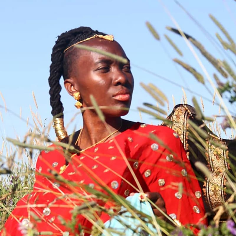 A woman in a red embellished dress and gold jewelry sits amidst tall grass against a clear blue sky.