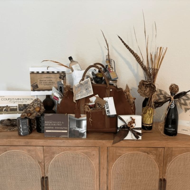 A collection of gift items, including a brown handbag, wine bottles, and gift certificates, sits atop a wooden sideboard against a plain white wall.