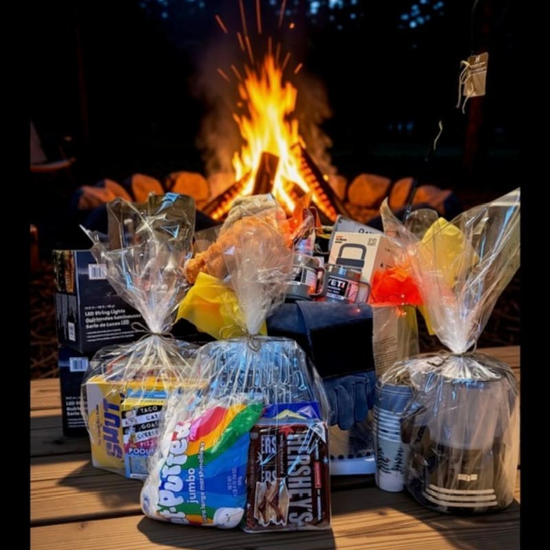 A collection of gift baskets filled with snacks and gear sits on a wooden table in front of a glowing campfire.