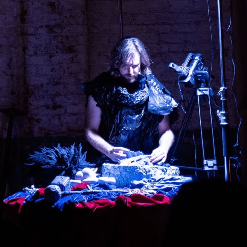 A man in a dark, textured garment stands over a table of various objects in a dimly lit room with a brick wall and camera equipment in the background.