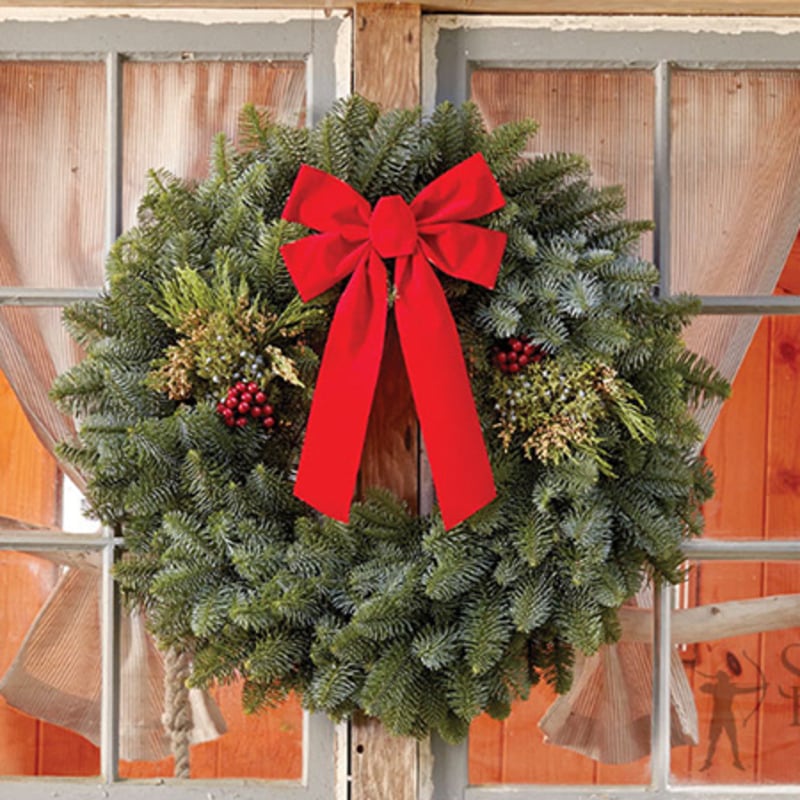 A lush green evergreen wreath with a large red bow and red berries hangs against a rustic wooden window frame.