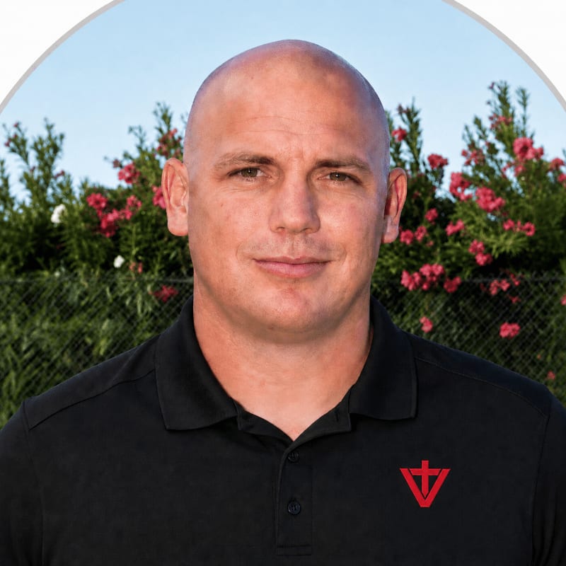 A bald man in a black polo shirt stands in the foreground against a background of green bushes with pink flowers.