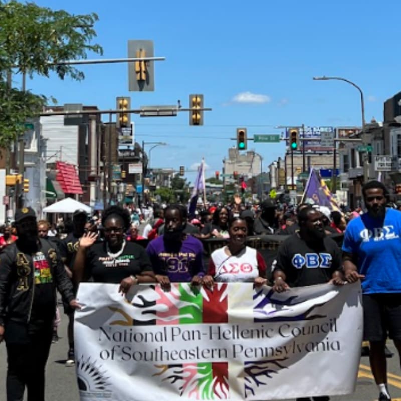A group of people hold a large white banner for the National Pan-Hellenic Council of Southeastern Pennsylvania on a sunny city street lined with buildings and traffic lights.