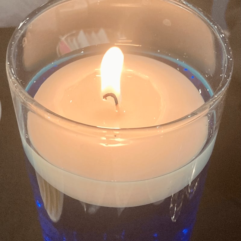 A lit white candle floats in a glass of blue liquid against a dark, blurred background.