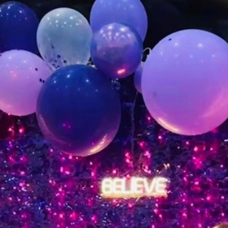 Purple and blue balloons float above a shimmering, glittery background featuring a glowing "BELIEVE" sign.