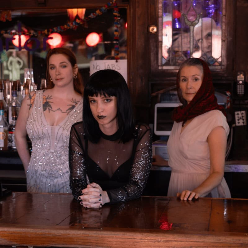 Three women stand behind a wooden bar counter in a dimly lit room with bottles and a mirror in the background.