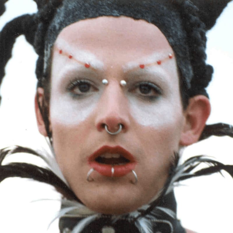 A person with white face paint, facial piercings, and a feathered collar is positioned against a plain, light-colored background.