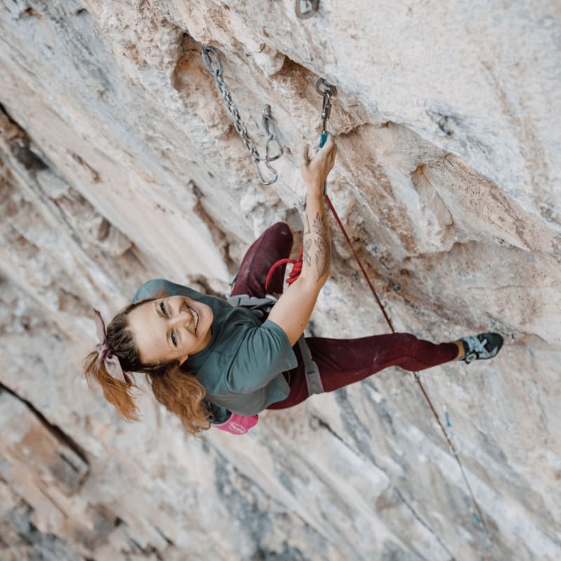 A smiling woman in climbing gear scales a light-colored, textured rock wall.