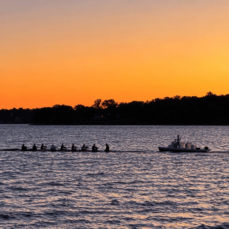 A rowing crew and a motorboat glide across the water in the foreground against a backdrop of silhouetted trees and a vibrant orange sunset.