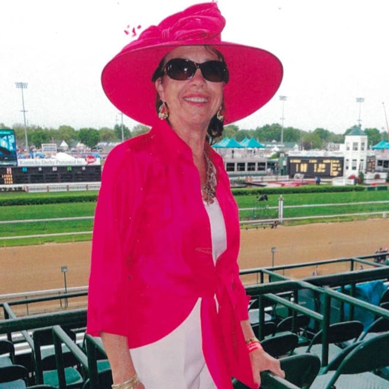 A woman wearing a bright pink hat and matching jacket stands in the foreground before a horse racing track and stadium seating in the background.