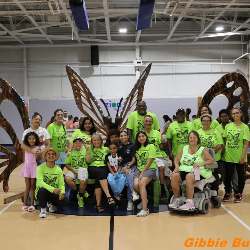 A group of people wearing bright green shirts pose together in the foreground of an indoor gymnasium featuring a large wooden butterfly sculpture in the background.