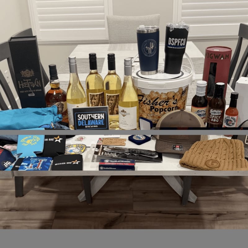 A variety of gift items, including wine bottles, tumblers, and apparel, are arranged on a white table in a brightly lit room.
