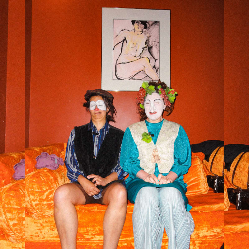 Two people in theatrical costumes and makeup sit on an orange sofa in front of a red wall featuring a framed sketch of a nude woman.