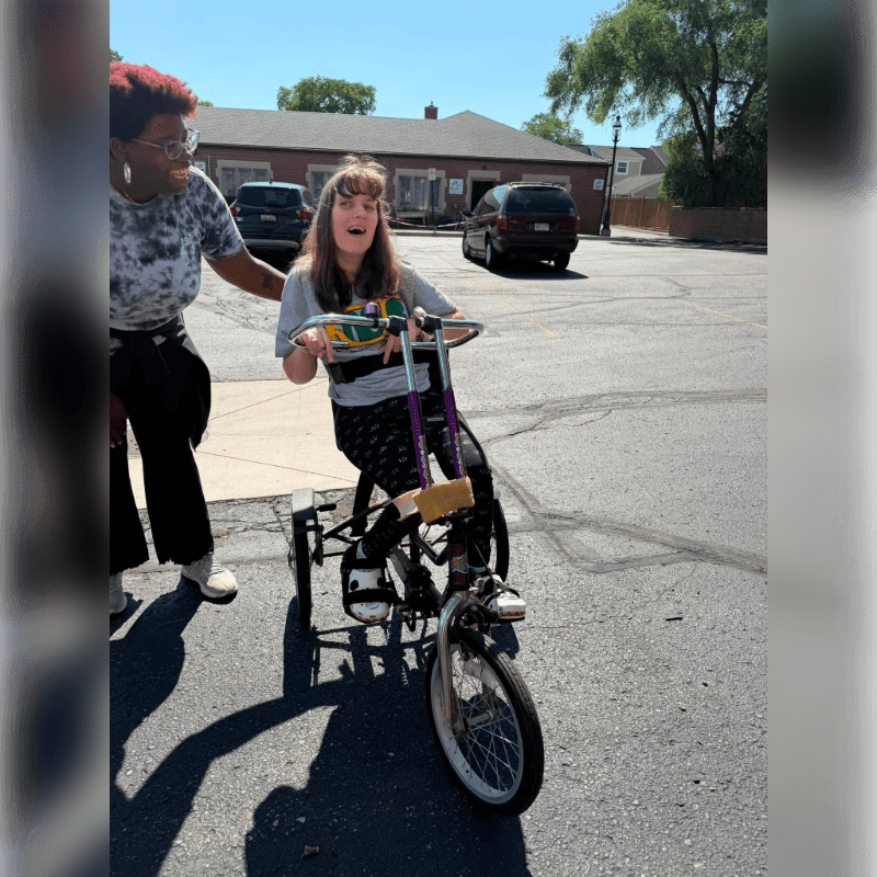 A woman with an excited expression rides a small bicycle while another woman stands beside her on a paved street with cars and a building in the background.