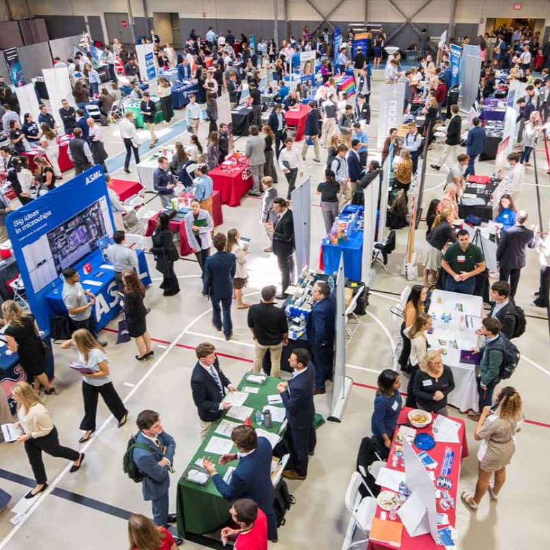 Numerous professionals and students mingle around various company booths and displays within a large, open exhibition hall.