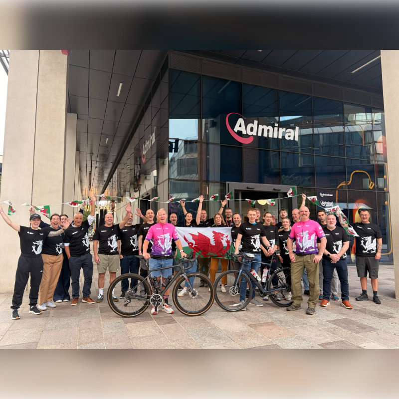 A group of people posing with bicycles and a Welsh flag stands in the foreground in front of a modern Admiral building.
