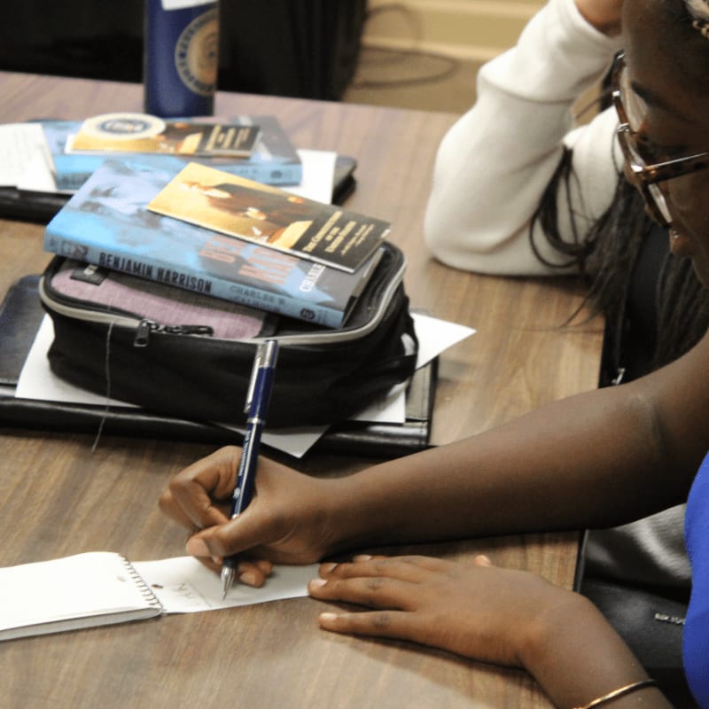 A person writes in a notebook on a wooden table, with books and a black bag resting in the background.