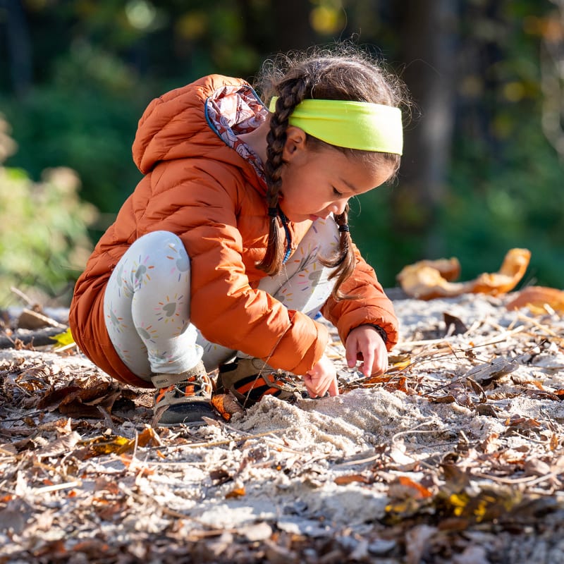 A young girl in an orange jacket and yellow headband crouches to play in the sand and leaves, with a blurred forest background.