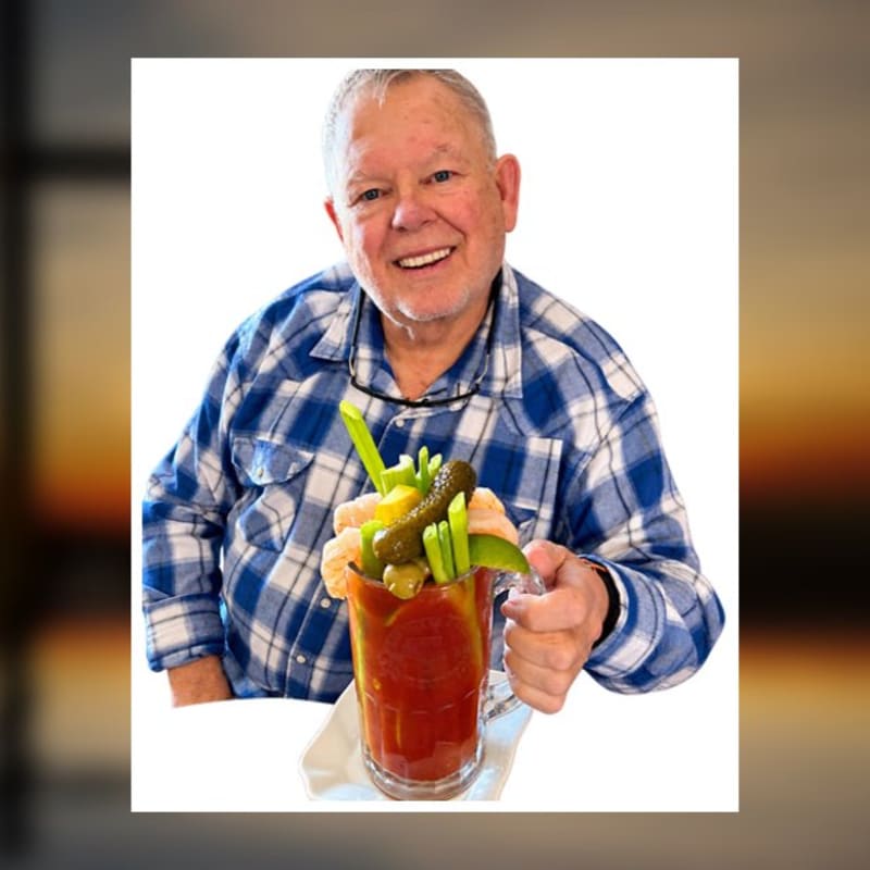 A smiling man in a blue plaid shirt holds a heavily garnished Bloody Mary against a plain white background.