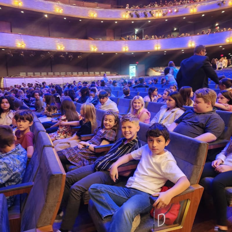 A group of children sit in theater seats in the foreground, while a large audience fills the auditorium and balconies in the background.