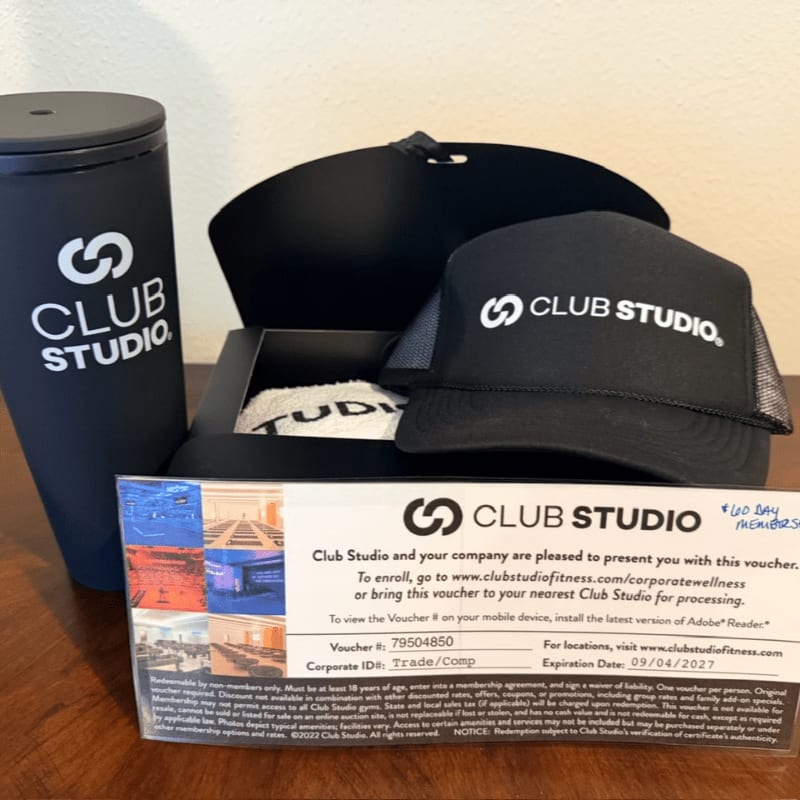 A black Club Studio tumbler, a matching baseball cap, and a membership voucher sit in the foreground against a plain, light-colored wall.