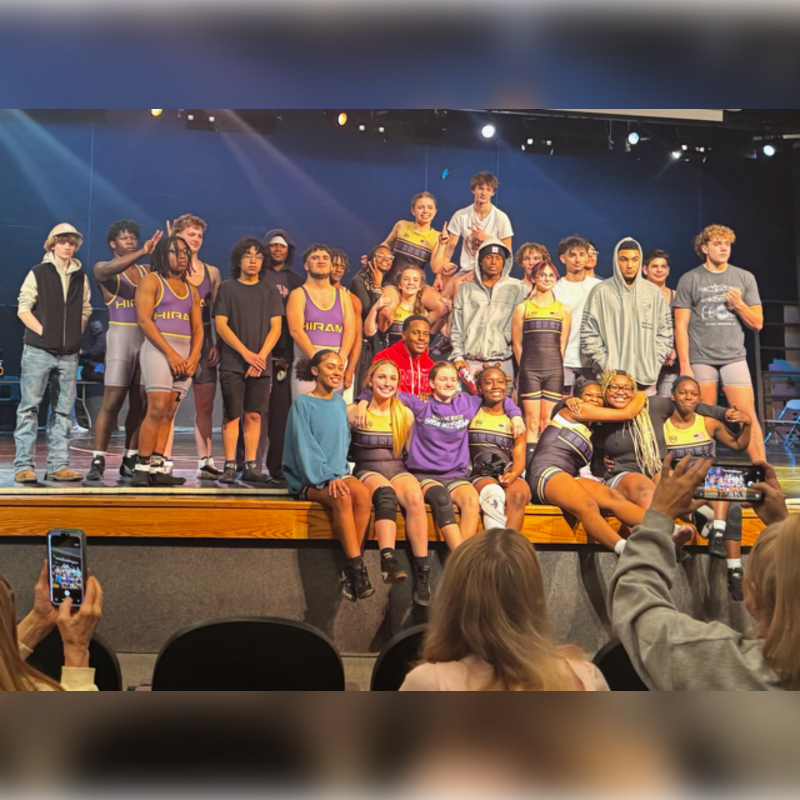A group of students in athletic wear poses for a photo on a stage, while people in the foreground capture the moment with their phones.