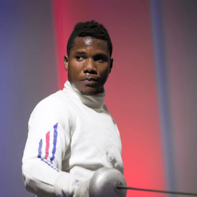 A fencer in white gear stands in the foreground against a blurred red and blue background.