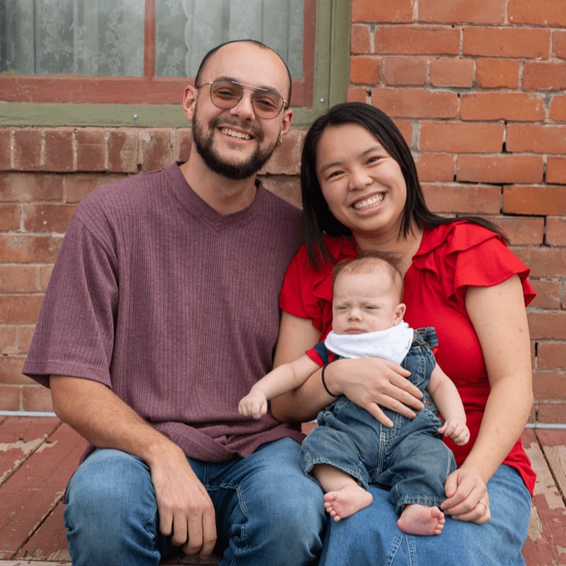 A smiling couple and their baby sit together in the foreground against a red brick wall background.