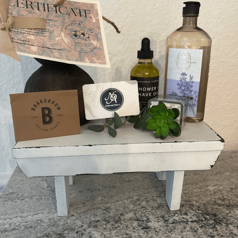 A gift set featuring skincare products, a gift certificate, and a small succulent sits on a white wooden riser against a neutral wall.