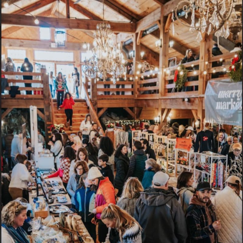 A crowd of people shops for goods in a rustic wooden market hall illuminated by ornate crystal chandeliers.