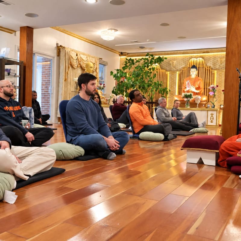 People sit cross-legged on cushions in a wooden-floored room, with a golden Buddha statue positioned against a decorative wall in the background.