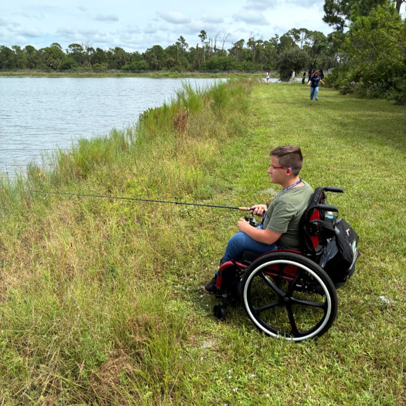 A boy in a wheelchair fishes from a grassy bank beside a calm lake with a line of trees in the distance.