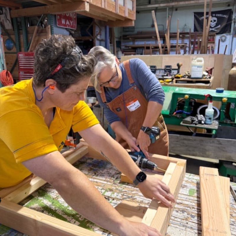 Two women work on a wooden frame in a cluttered woodworking shop filled with tools and equipment.