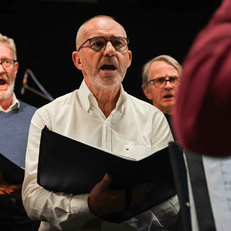 A man in a white shirt sings while holding a black folder, flanked by other male singers against a dark background.