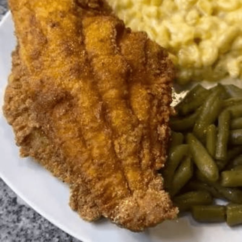 A piece of golden-brown fried fish sits in the foreground, accompanied by macaroni and cheese and green beans in the background.