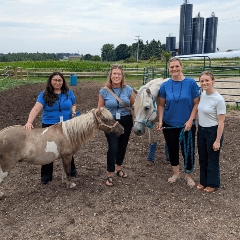 Four women stand with two miniature horses in a dirt paddock, with tall silos and green trees in the background.