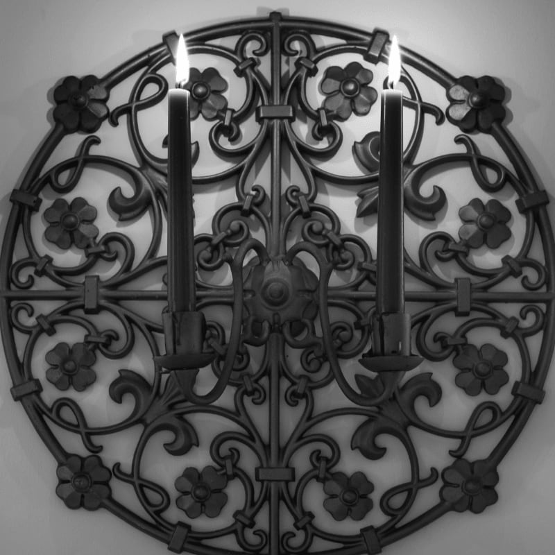Two lit black candles stand in the foreground against a circular, ornate wrought-iron floral backdrop on a plain wall.