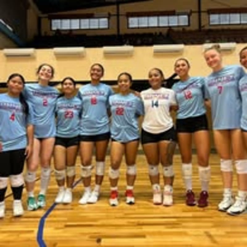 A volleyball team in light blue and white uniforms poses together on a polished wooden gym floor against a background of indoor stadium seating and windows.