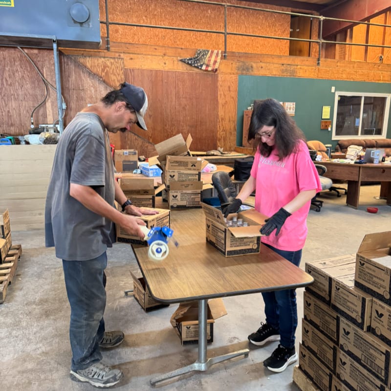 A man and a woman pack boxes on a table inside a rustic wooden warehouse.