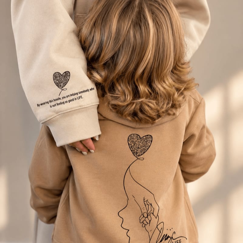 A person with wavy blonde hair wears a tan hoodie featuring heart graphics and text, set against a soft, blurred neutral background.