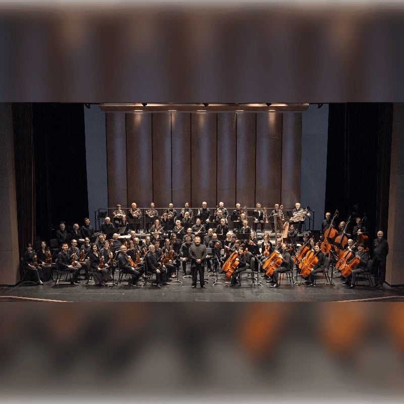 An orchestra of musicians with their instruments sits on a stage in front of a tall, paneled wooden wall.