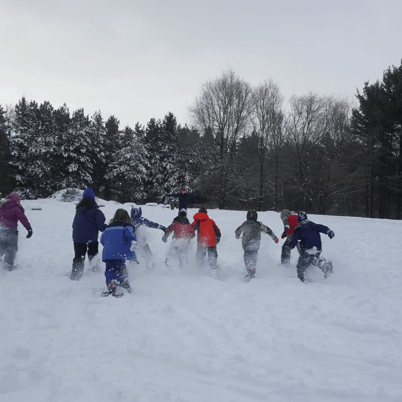 A group of children in colorful winter gear run through deep snow toward a backdrop of snow-covered evergreen and bare trees.